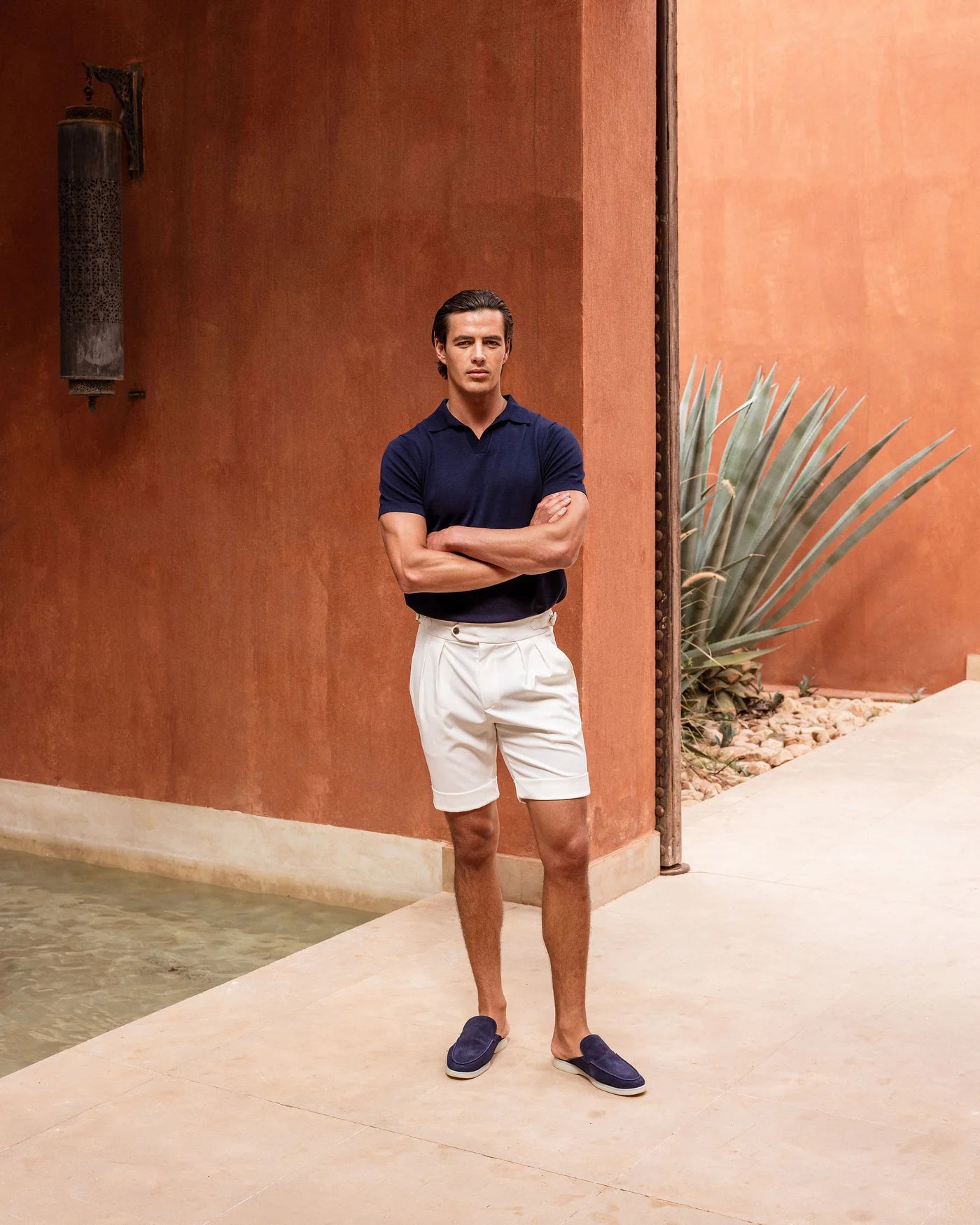 Man standing with arms crossed against a warm-toned wall with a plant.