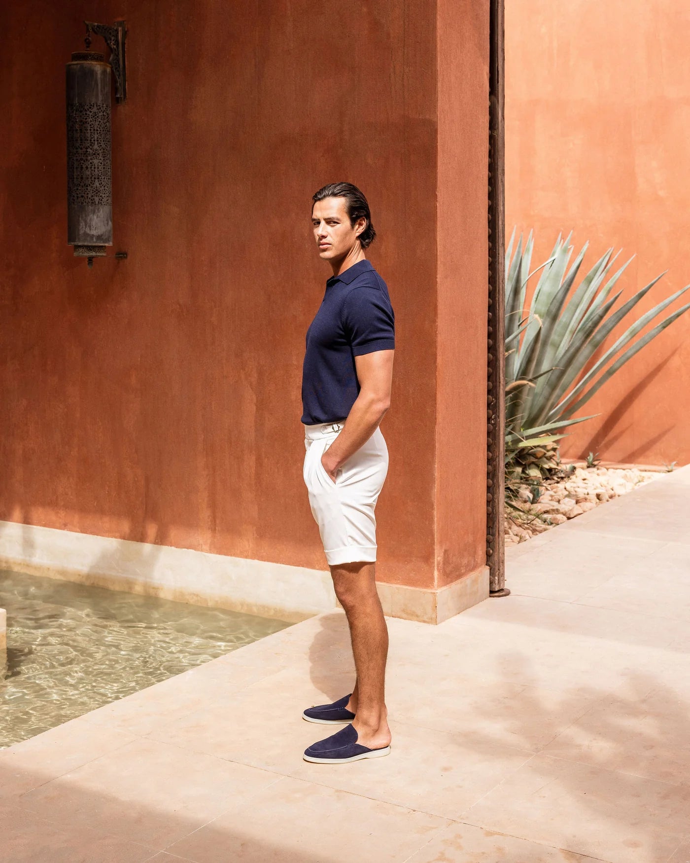 Man in navy shirt and white shorts standing against a red wall with a plant in the background