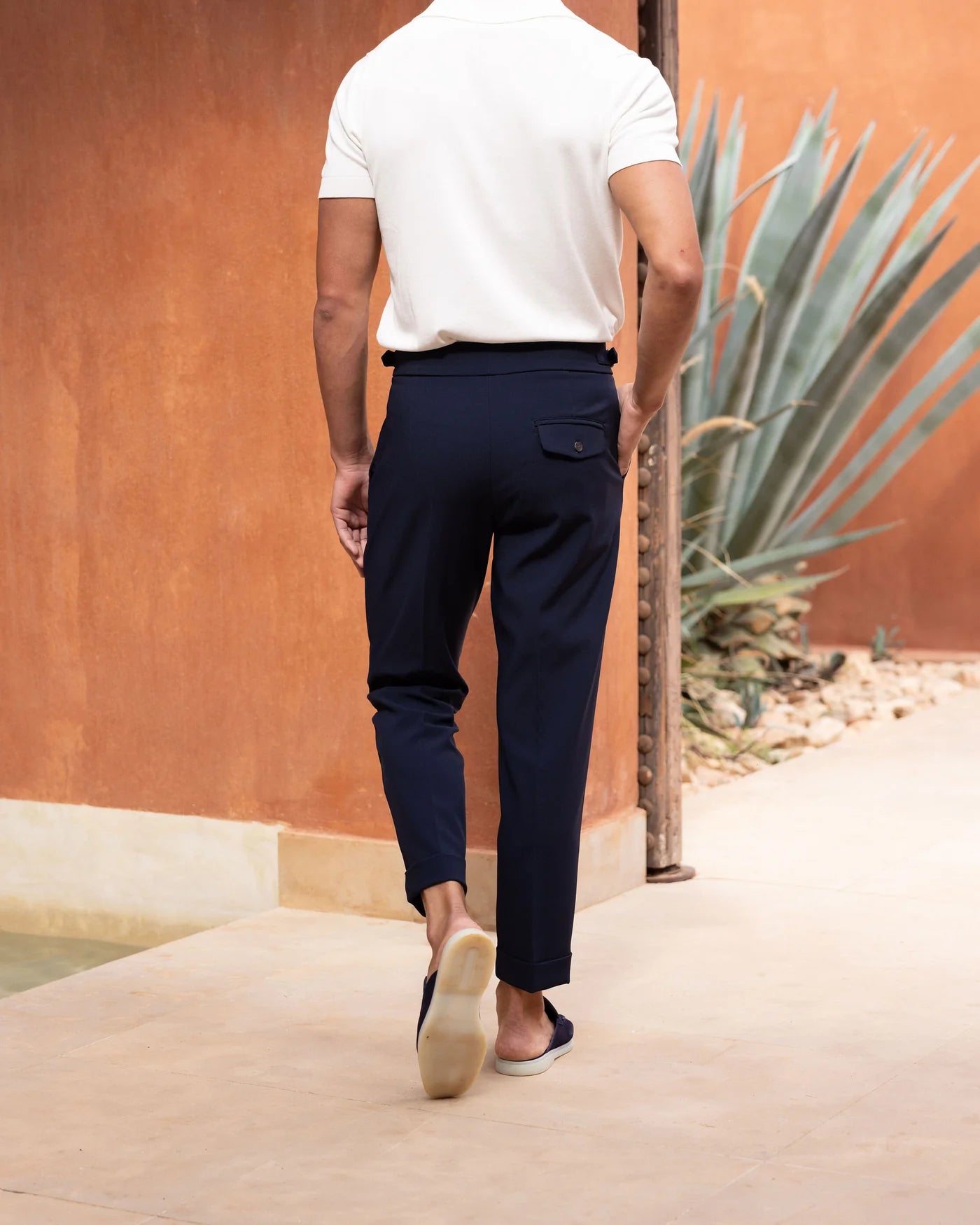 Person wearing navy pants and a white shirt standing against an orange wall with a plant.