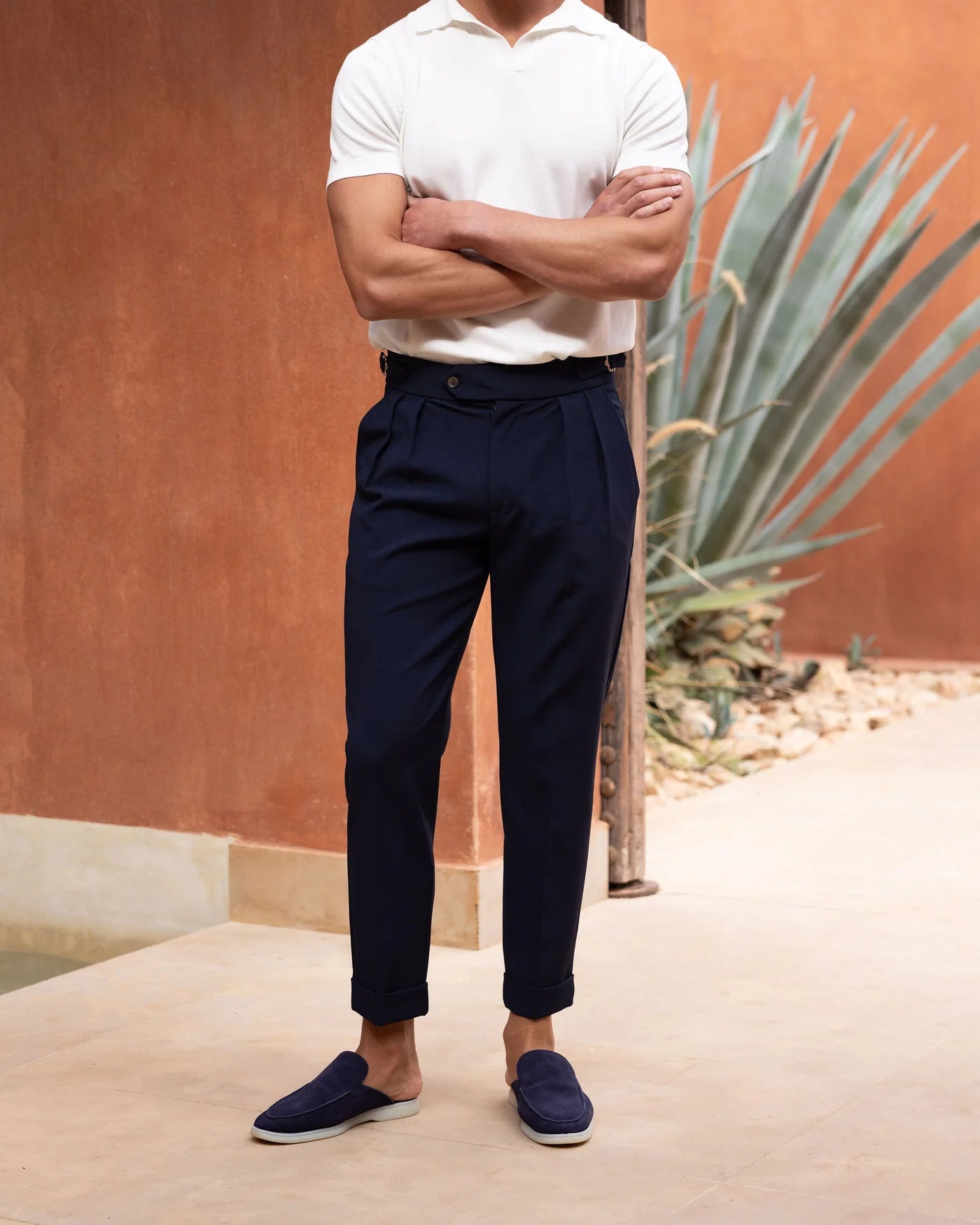 Man wearing a white polo shirt, navy pants, and blue shoes standing against a warm-toned wall with a plant.