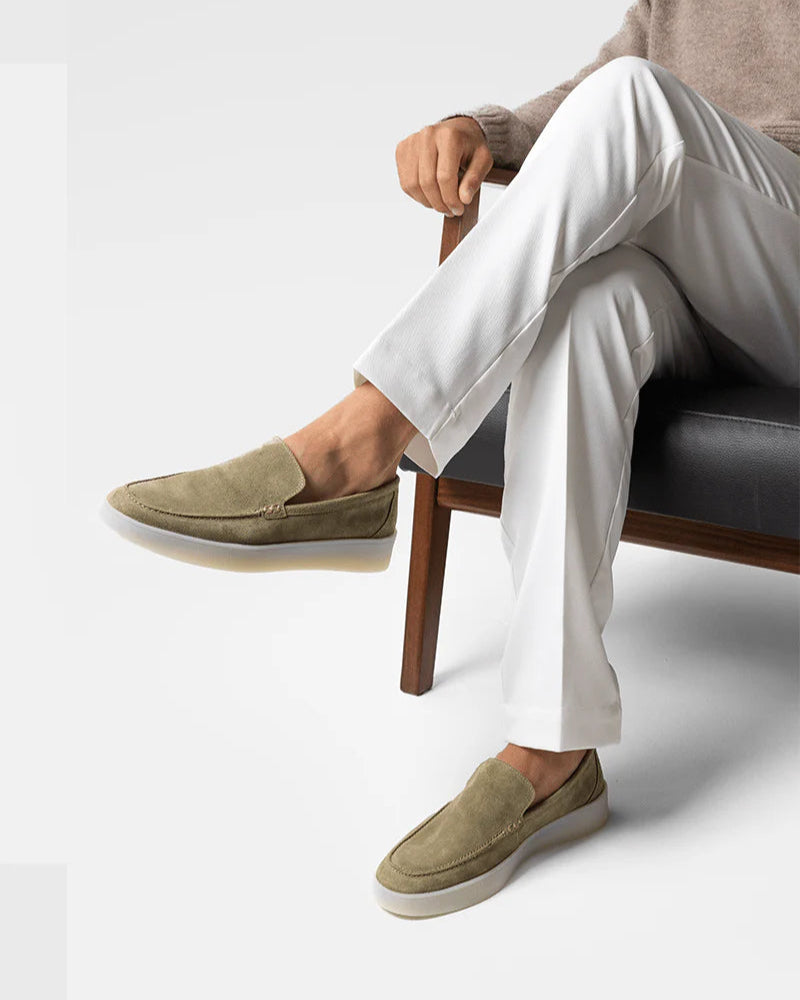 Person wearing beige loafers and white pants sitting on a chair with a plain background