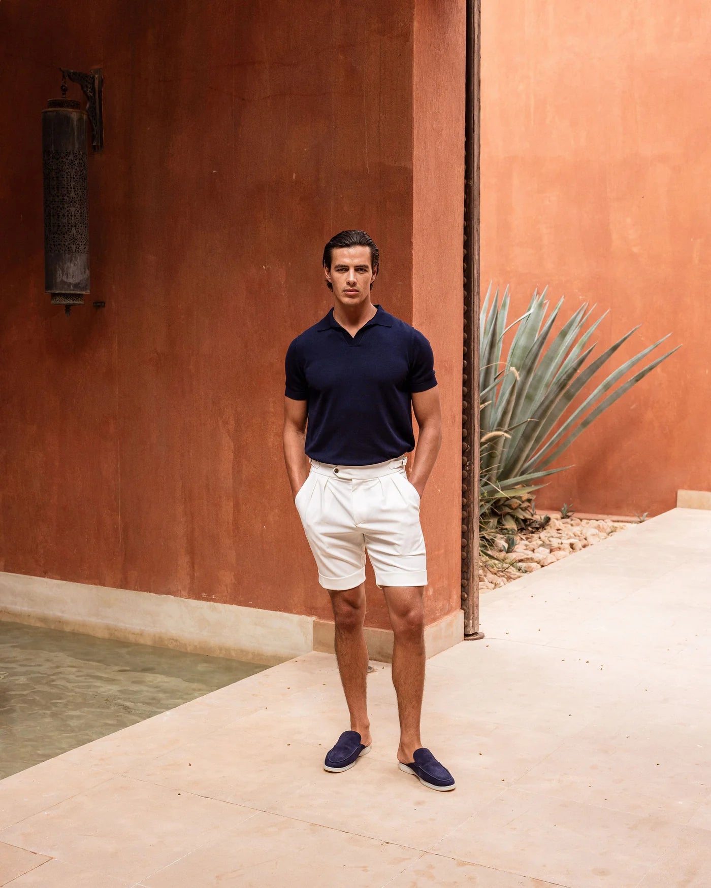 Man wearing a navy polo shirt and white shorts standing against a terracotta wall with a cactus plant.