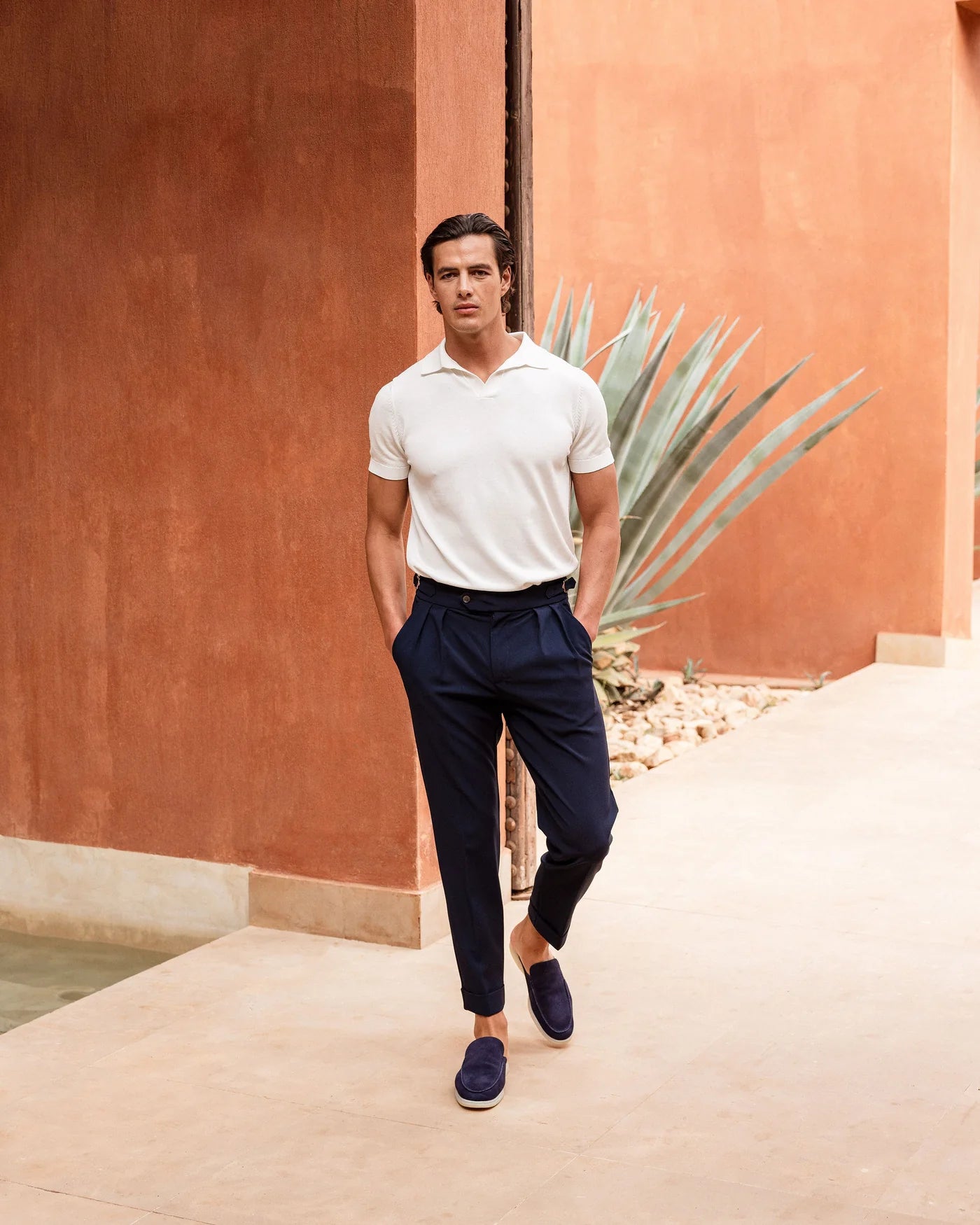 Man in a white polo shirt and navy pants standing against a warm-toned wall with a plant.